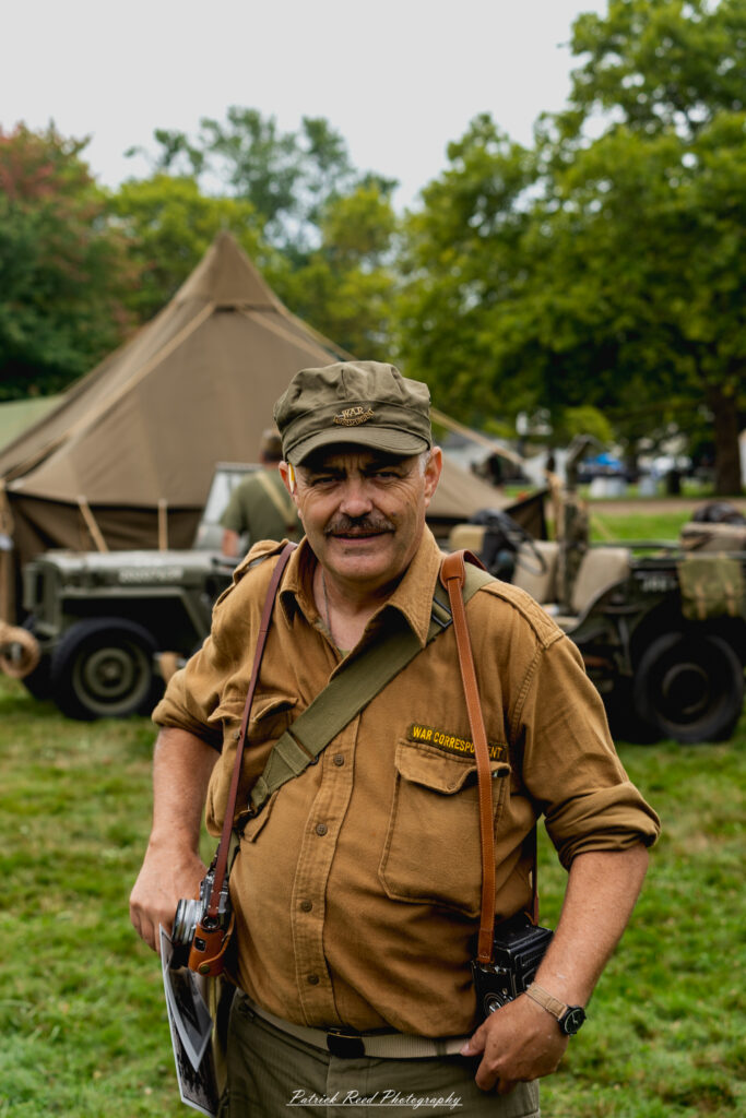 "A World War II war correspondent, dressed in a rugged, practical outfit suitable for the battlefield. Equipped with a camera slung over one shoulder and a notepad in hand, the correspondent captures the intense realities of war. Their helmet and press badge mark them as a non-combatant, while their expression reflects a mix of determination and focus as they report from the front lines. The background includes war-torn surroundings, hinting at the dangers and challenges of documenting history in real time."